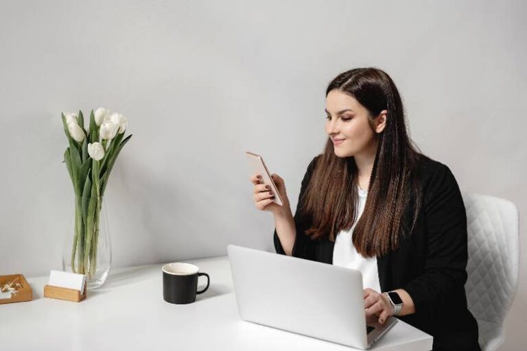 Woman sitting at a desk with a laptop, looking at her phone - Facebook groups for women entrepreneurs.