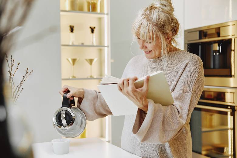 Woman in a kitchen, reading a book while pouring water into a teacup - how to reinvent yourself at 40.