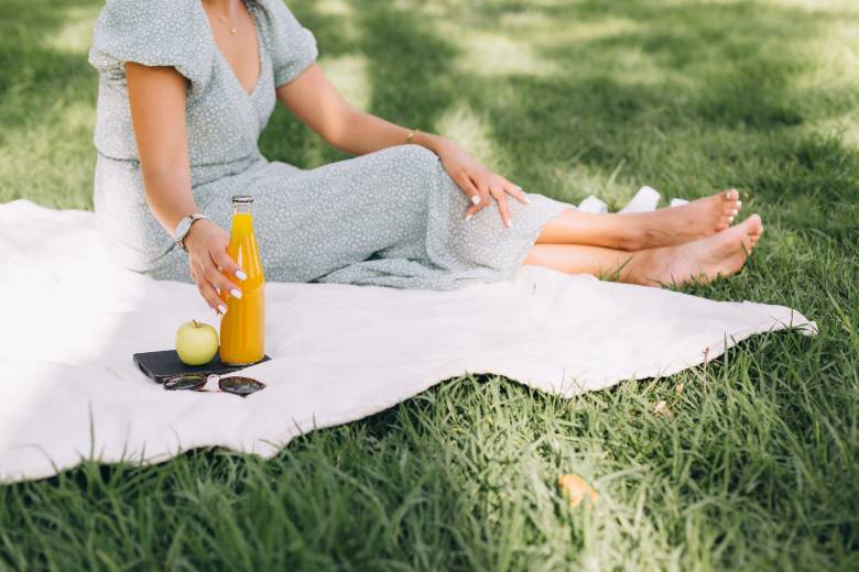 Faceless woman enjoying a drink and snack on a blanket on grass - Spring self-care ideas.