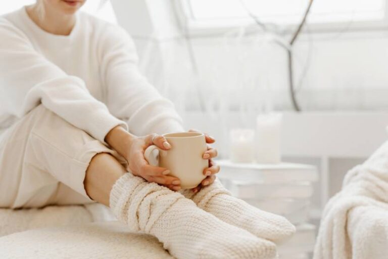 Faceless woman in a cozy white sweater and socks, sitting holding a cup - winter activity ideas.