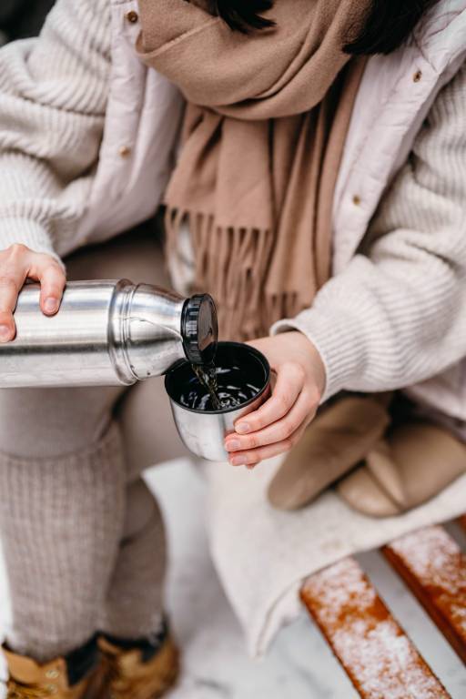 Faceless woman pouring hot coffee into a thermos mug while wearing winter clothes - winter activity ideas.